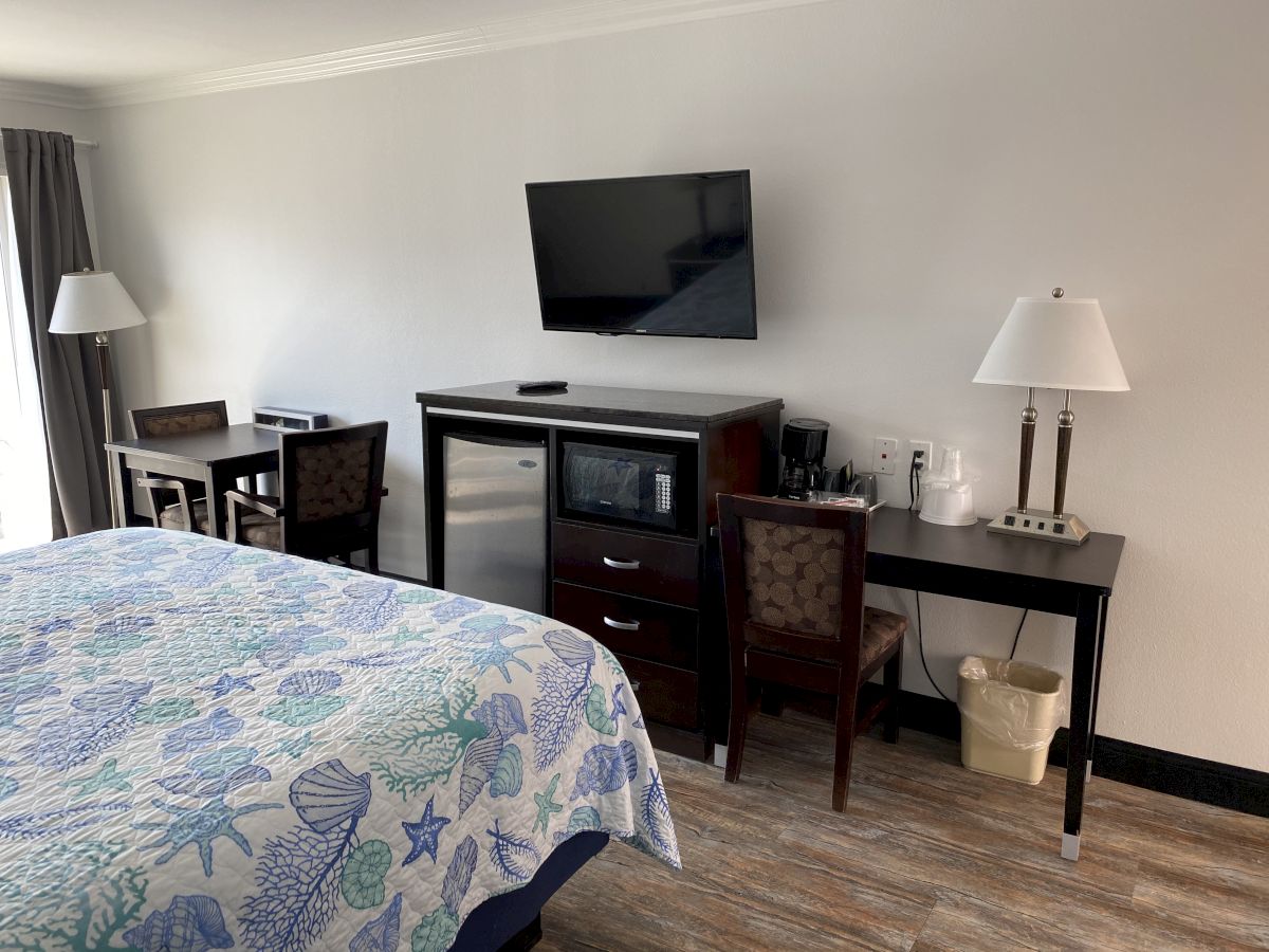 The image shows a hotel room featuring a bed with a sea-themed quilt, two lamps, a wall-mounted TV, a microwave, and a desk with chairs.