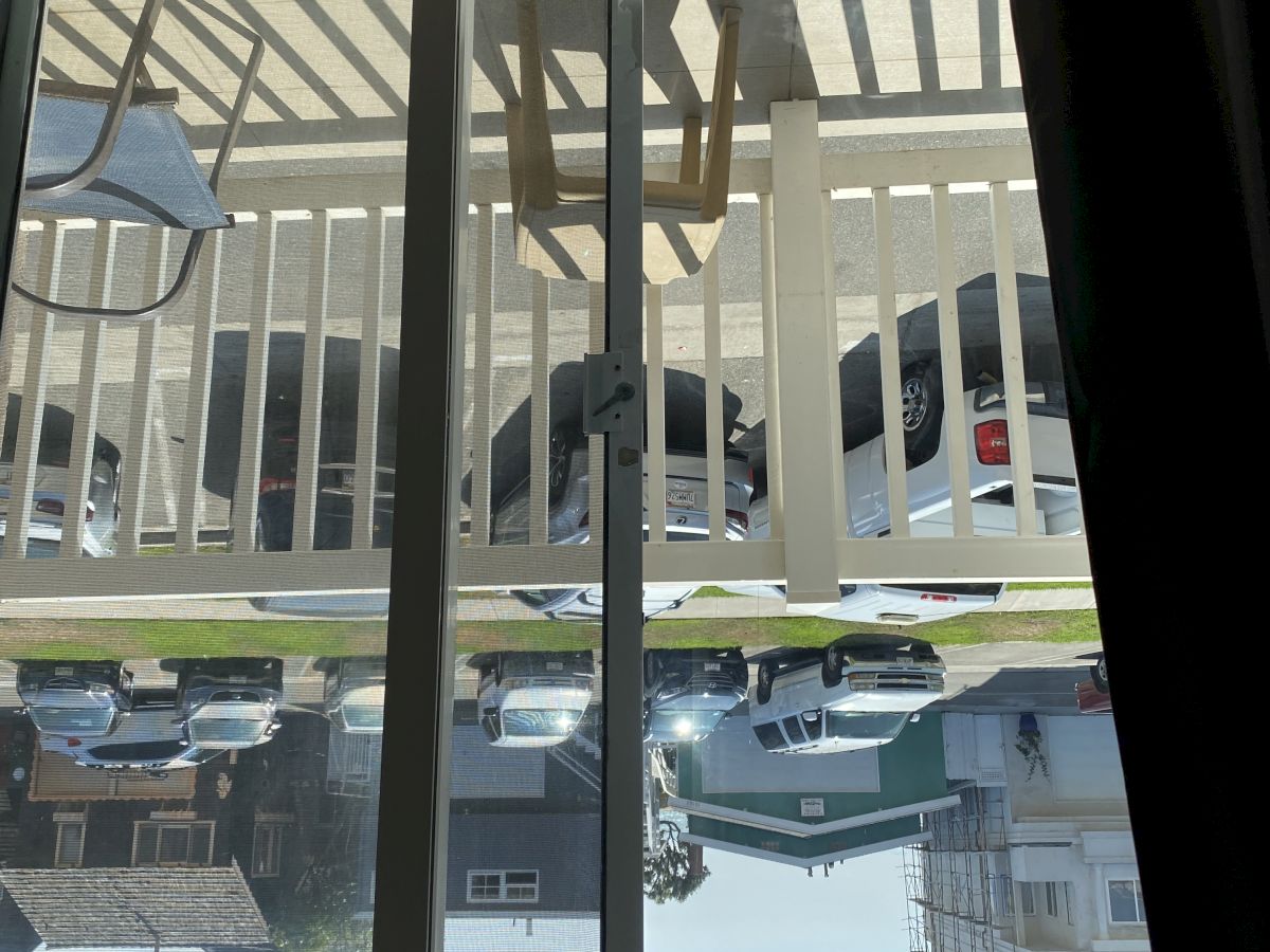 The image shows a view through glass doors leading to a balcony with a chair and table, overlooking parked cars and a residential street.