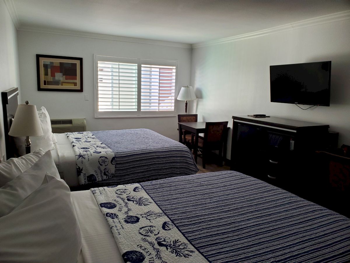 A neatly arranged hotel room with two beds, wall art, a window, a desk with a lamp, and a wall-mounted TV opposite the beds.