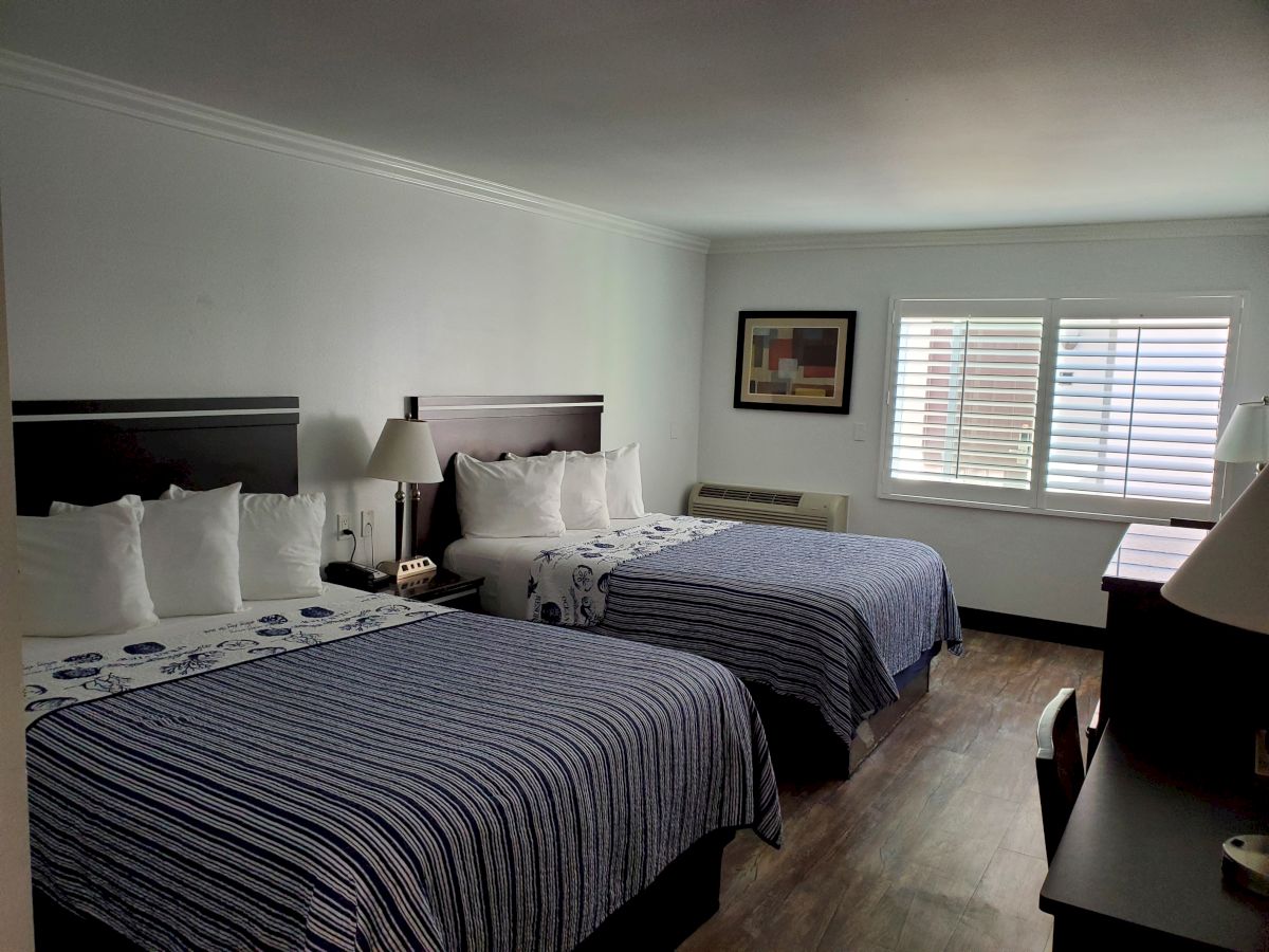 A hotel room with two double beds, white pillows, striped bed covers, a desk, bedside lamps, a wall painting, and a window with blinds.