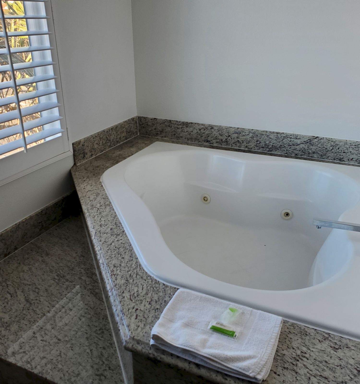 A clean white bathtub is set in a granite counter next to a window with blinds. A towel and soap bar are placed on the counter.