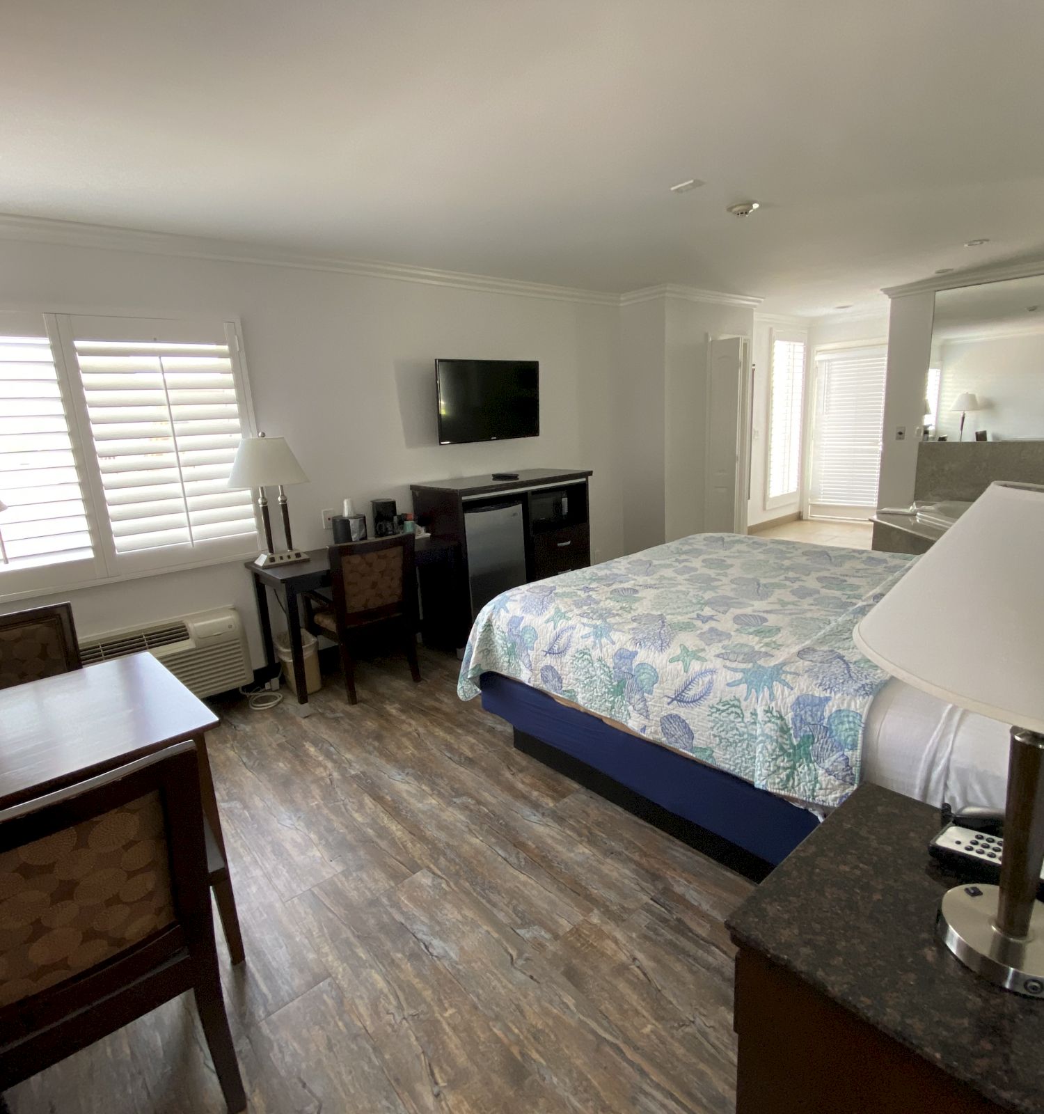 A hotel room with a bed, two chairs, two lamps, a desk, a dresser, a flat-screen TV, and wooden flooring. There's a window with shutters.