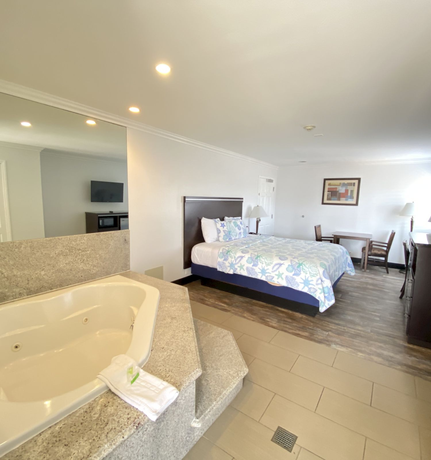 This image shows a hotel room with a large bed, a jacuzzi tub, a small desk, a chair, a TV mounted on the wall, and a picture on the wall.