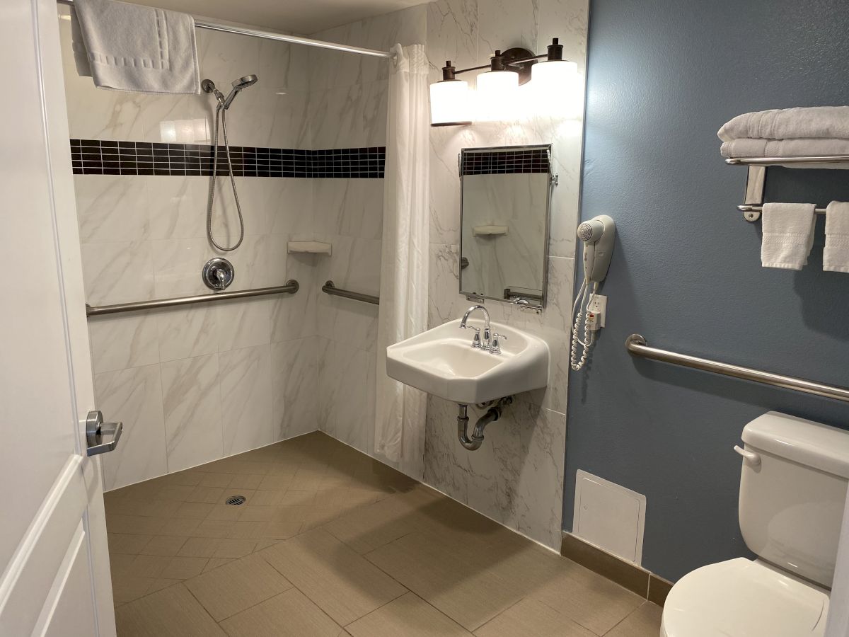 This image shows an accessible bathroom with a walk-in shower, grab bars, a sink, a toilet, a towel rack, and a mirror, providing a safe environment.