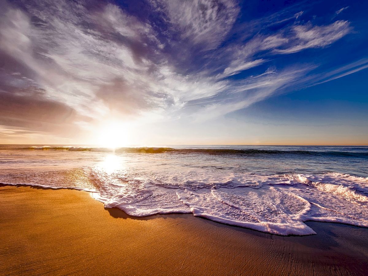 An ocean sunset with golden sandy shores and waves lapping the beach, under a partly cloudy sky with hues of blue, orange, and purple ending the sentence.