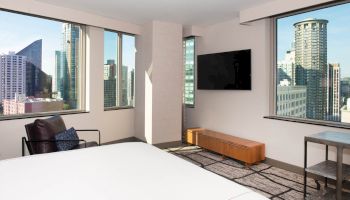 A modern hotel room with a city view, a bed, a chair, a wall-mounted TV, and large windows letting in natural light.
