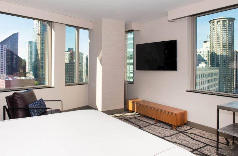 A modern hotel room with a city view, a bed, a chair, a wall-mounted TV, and large windows letting in natural light.