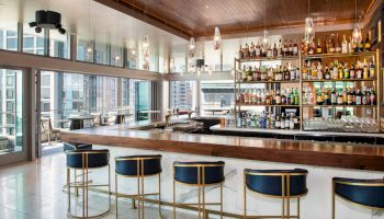 A modern bar with high ceilings, stylish hanging lights, bottles of alcohol on shelves, and a long counter with stools, set in a high-rise building.