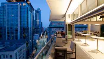A modern rooftop terrace with seating and tables, overlooking a city street lined with tall buildings at dusk, creating a cozy urban atmosphere.
