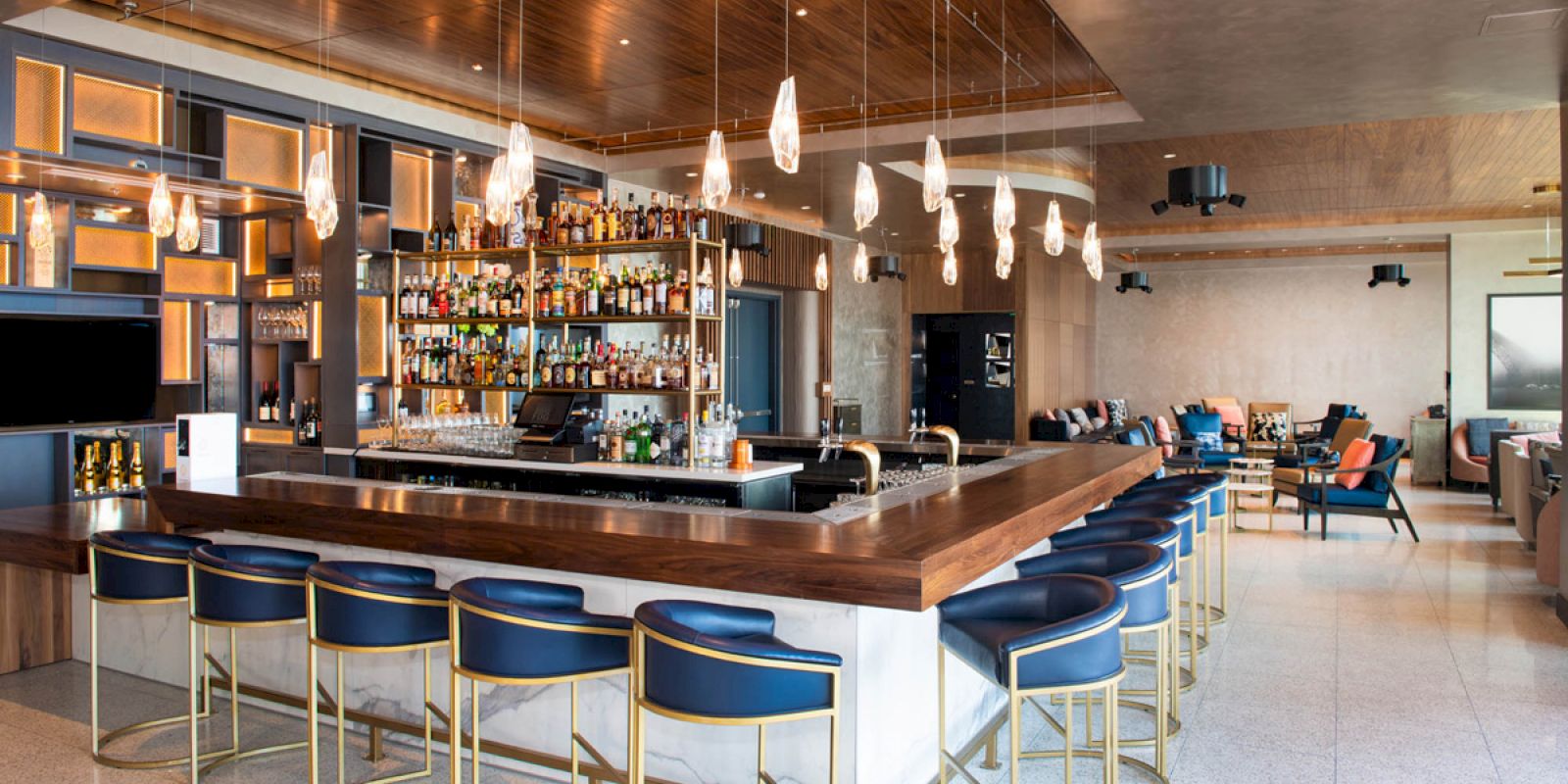 The image depicts a modern bar with a wooden counter, blue bar stools, hanging pendant lights, and a well-stocked shelf of beverages in the background.