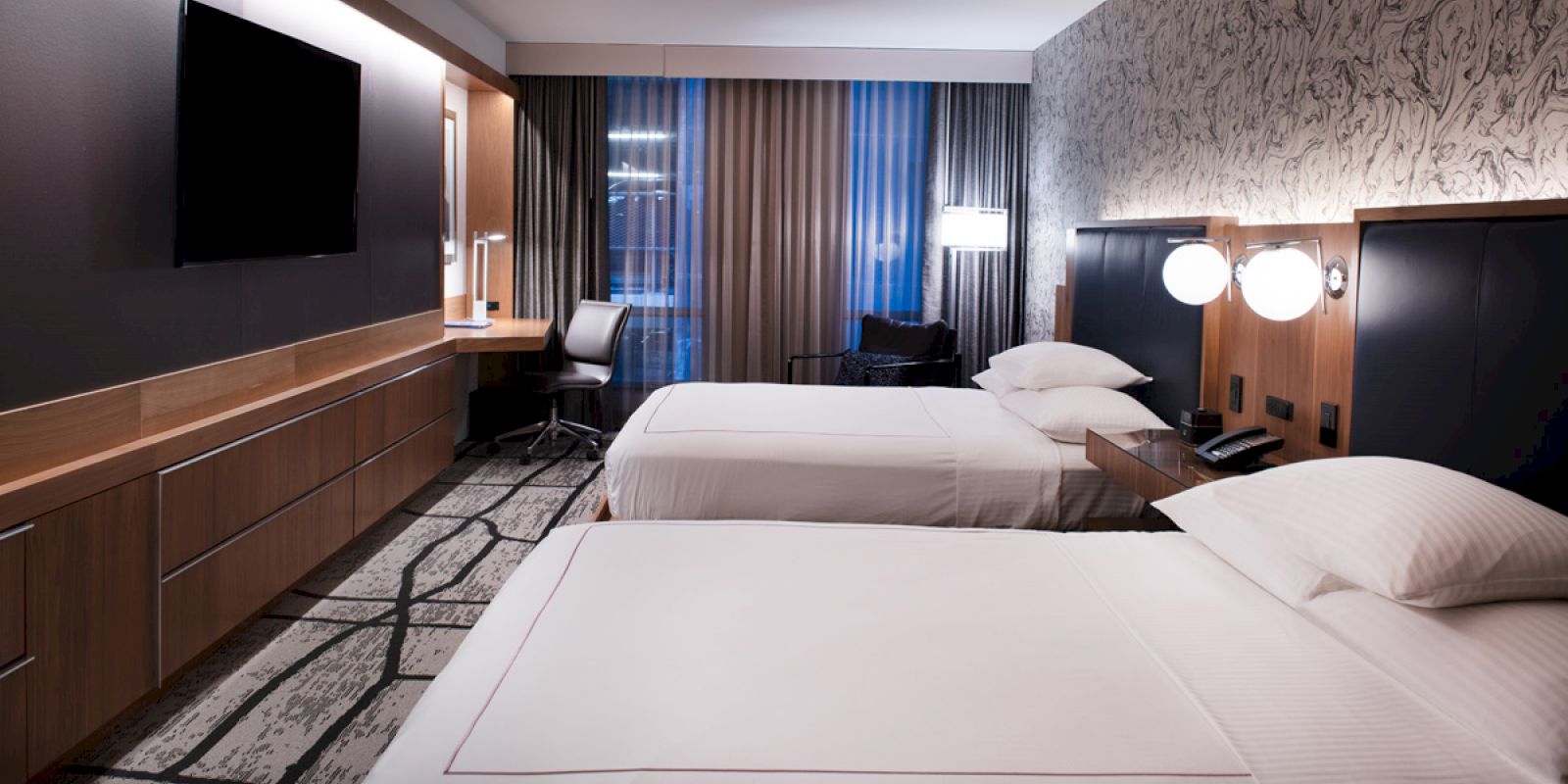 A modern hotel room with two beds, a large TV, a desk and chair near the window, and contemporary lighting and decor on the walls ending the sentence.