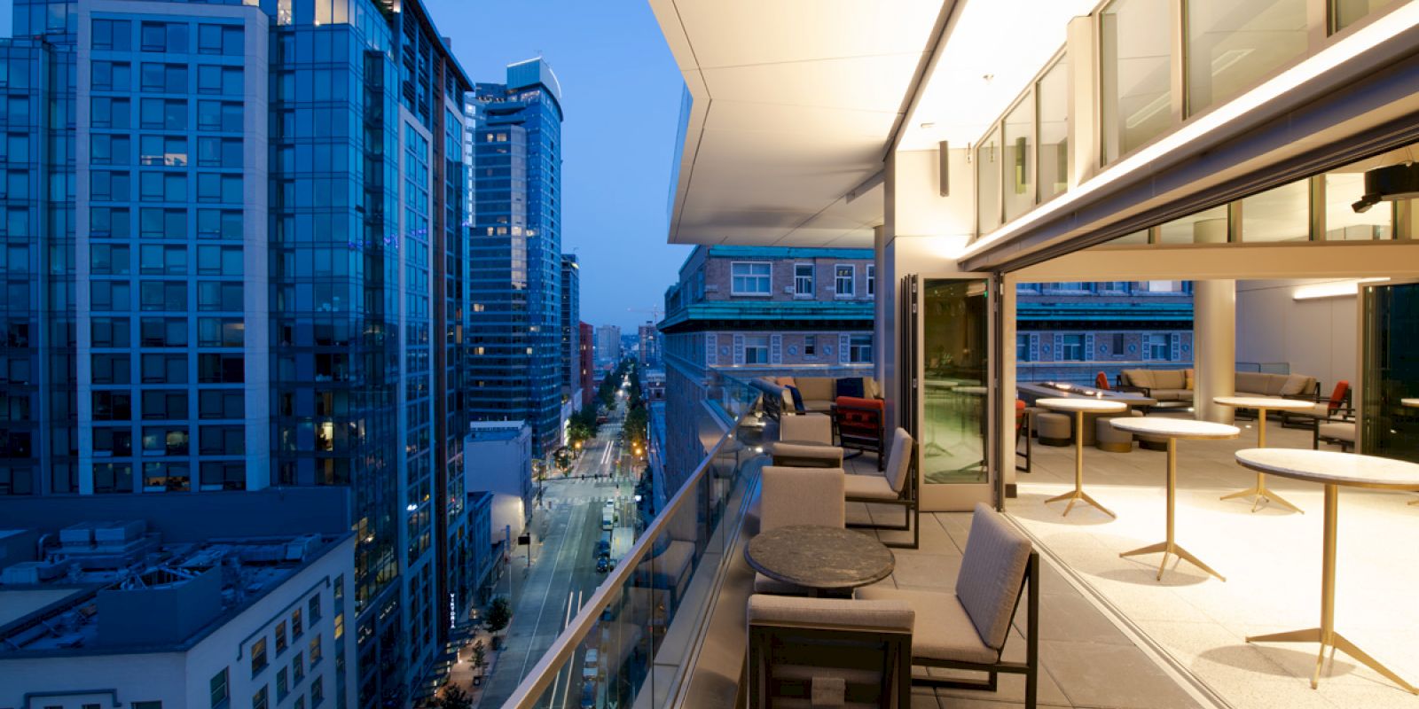 An outdoor terrace at dusk with seating, overlooking a cityscape featuring modern high-rise buildings and well-lit streets.