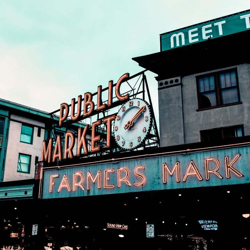 The image shows the famous Public Market sign at Pike Place Market in Seattle, with a clock and Farmers Market signage prominently displayed.