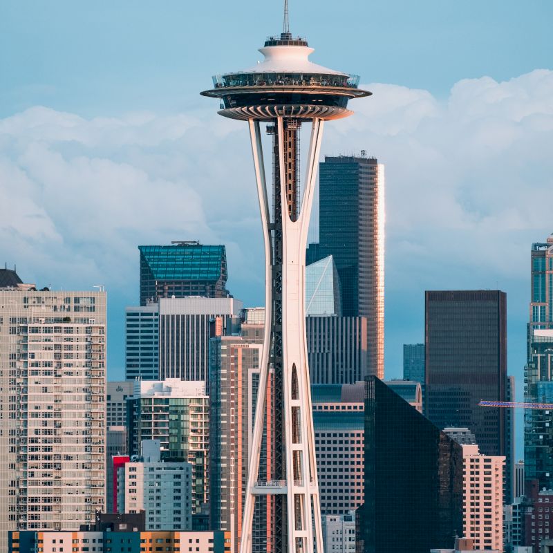 The image shows the iconic Space Needle tower with a backdrop of the Seattle city skyline and various buildings under a partly cloudy sky.