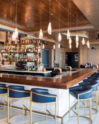 A modern bar with elegant lighting, high chairs, and a wide selection of bottles on display. There are lounge seating areas in the background.
