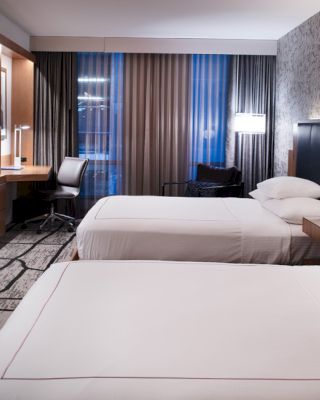 A modern hotel room with two double beds, wall-mounted TV, work desk, floor-to-ceiling windows with curtains, and stylish decor.