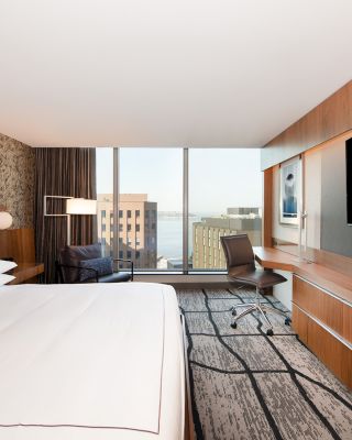 The image shows a modern hotel room with a large bed, desk, chair, TV, and floor-to-ceiling windows with a cityscape view and art on the wall.