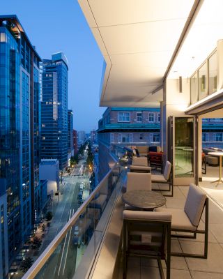 A rooftop patio with modern outdoor seating overlooks a city street and tall buildings at dusk, showcasing an urban and stylish setting.