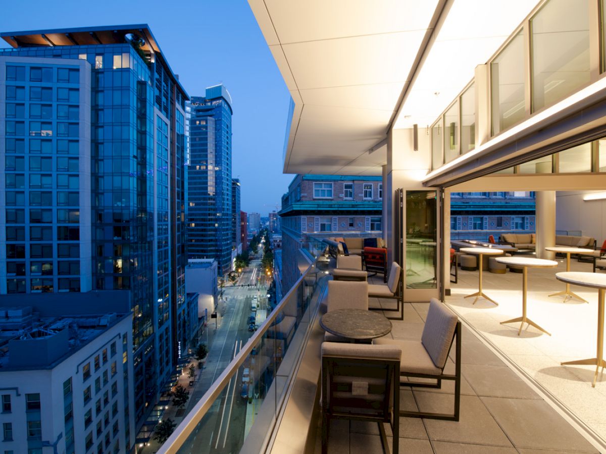 A modern balcony with seating and tables overlooks a city street lined with tall buildings, captured during twilight hours.