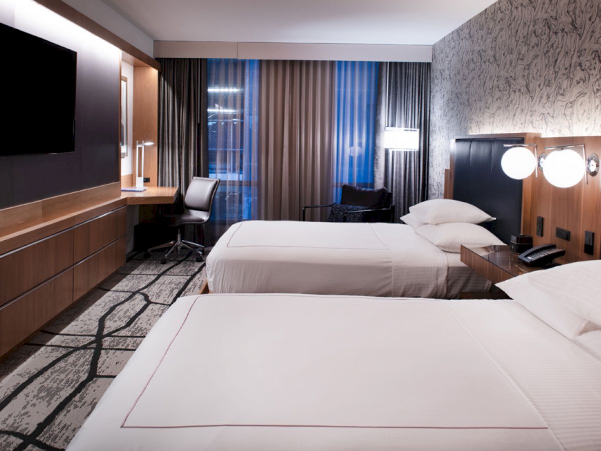 A modern hotel room features two neatly made beds, a desk with a chair, a flat-screen TV, and a patterned carpet and wall design.