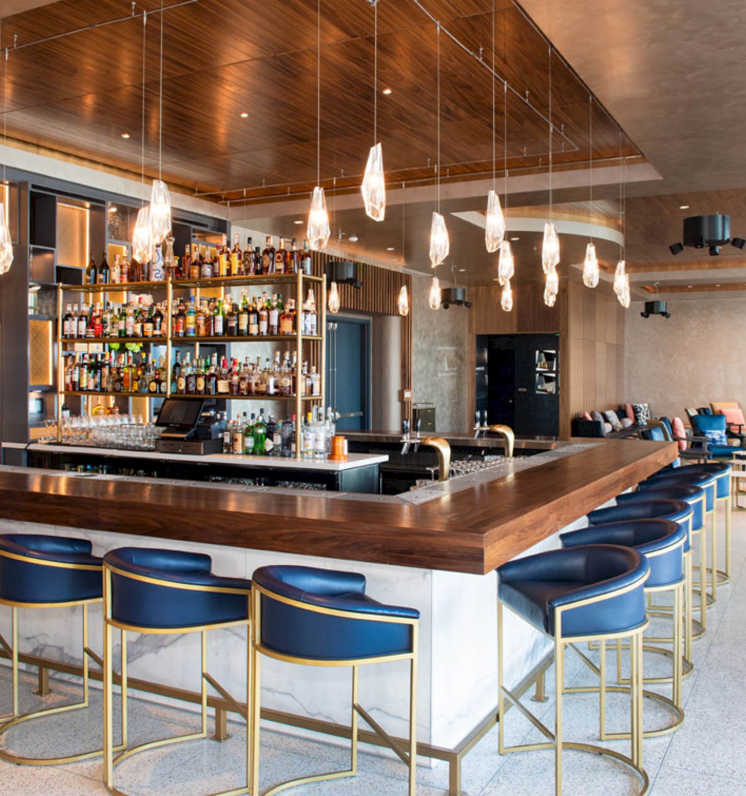 The image shows a modern bar with a central counter, blue bar stools, hanging lights, and a well-stocked back bar. The setting is elegant and inviting.