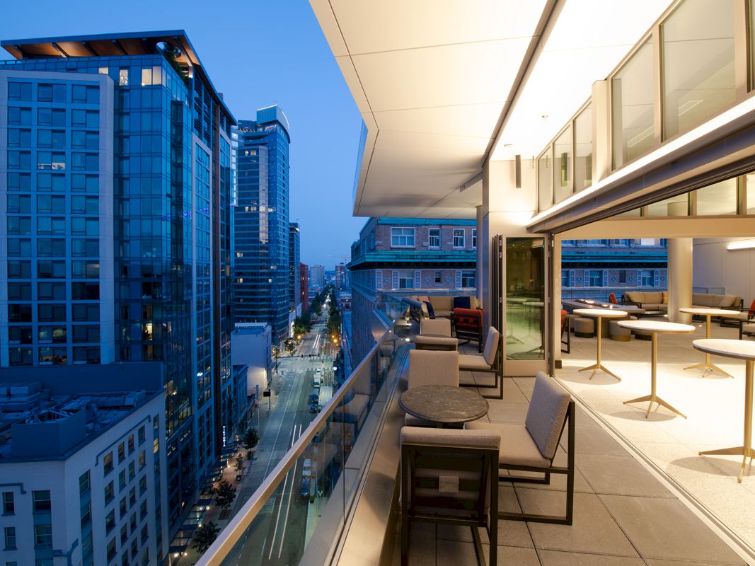 This image shows a modern cityscape view from a balcony during twilight, with buildings lining the street, and outdoor seating on the balcony.