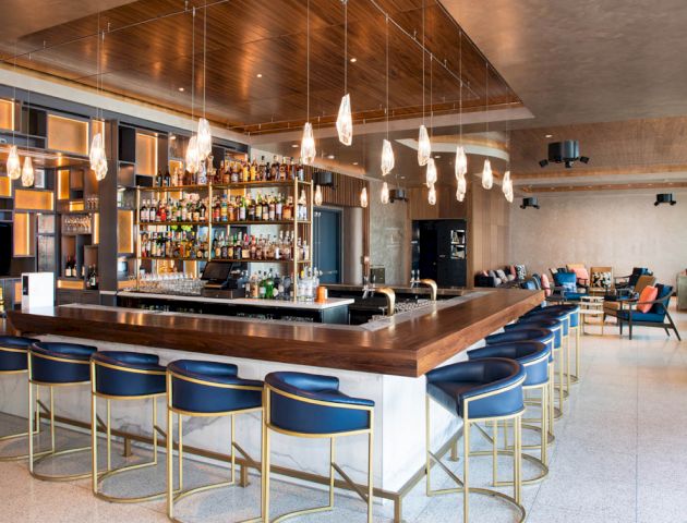 This image shows a modern bar interior with a wooden counter, blue barstools, a well-stocked bar, hanging lights, and a lounge area in the background.
