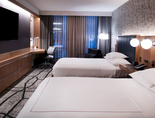 A modern hotel room with two beds, a wall-mounted TV, a desk with a chair, and floor-to-ceiling windows with curtains drawn.