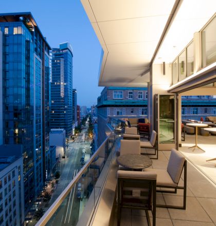 A modern cityscape at dusk with tall buildings. To the side, there’s a terrace with tables, chairs, and bright lighting.