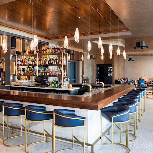 The image shows a modern bar with a large counter, multiple bar stools, hanging lights, and a well-stocked shelf of alcohol bottles.