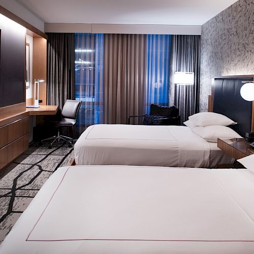 A modern hotel room with two beds, a flat-screen TV, a desk with a chair, and a window with curtains.