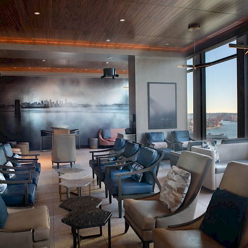 A modern lounge with comfortable seating, a cityscape mural, large windows with a waterfront view, and natural lighting filling the space.