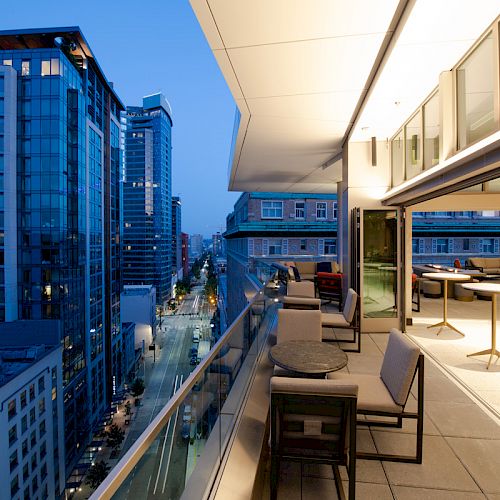 This image shows an urban balcony with modern outdoor seating, overlooking a cityscape with tall, well-lit buildings and a street below.