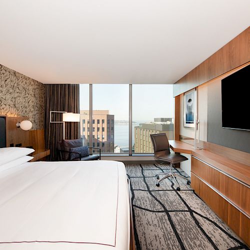 This image shows a modern hotel room with a large bed, a desk and chair, a flatscreen TV, and a large window offering a cityscape view.