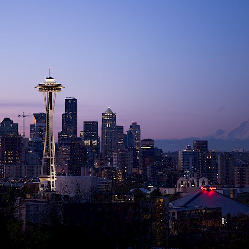 This image shows the Seattle skyline at dusk, featuring the iconic Space Needle with Mount Rainier visible in the background and city lights illuminating.