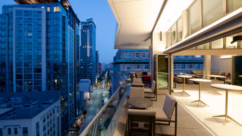 The image shows a modern balcony with seating and tables, overlooking a cityscape of tall buildings and a lit street at dusk.