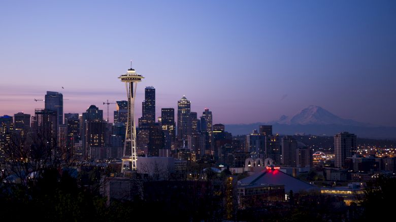 A nighttime view of a city skyline featuring the Space Needle, with a range of mountains visible in the background.