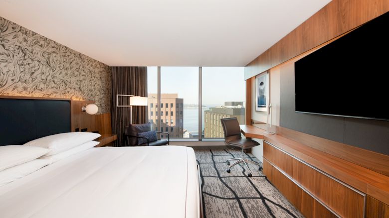 A modern hotel room with a large bed, a desk, a TV, and a window with a city view. The decor is sleek with wooden accents and neutral tones.