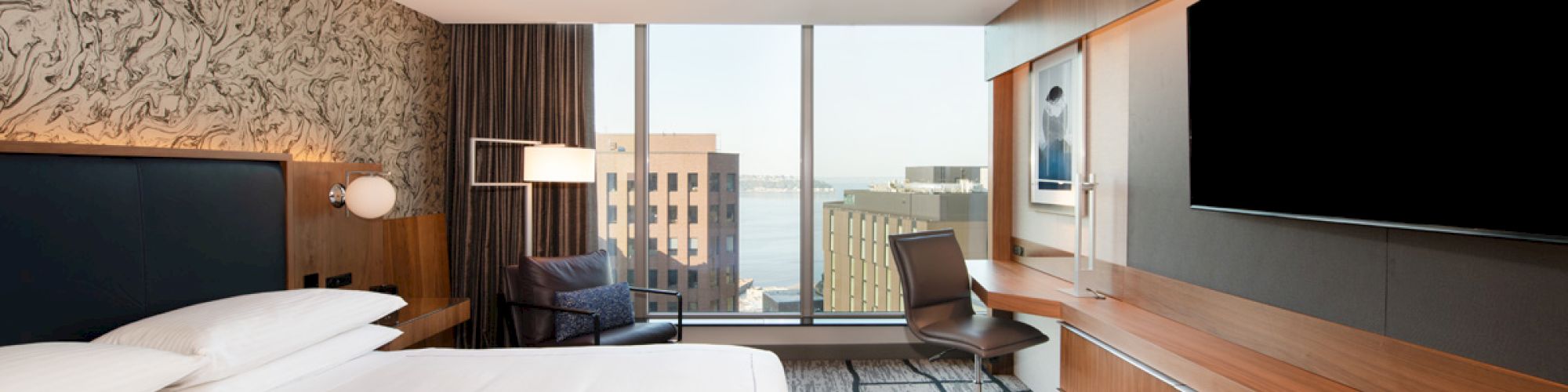 A modern hotel room with a large bed, a wall-mounted TV, a desk, and a view of city buildings and water through floor-to-ceiling windows.