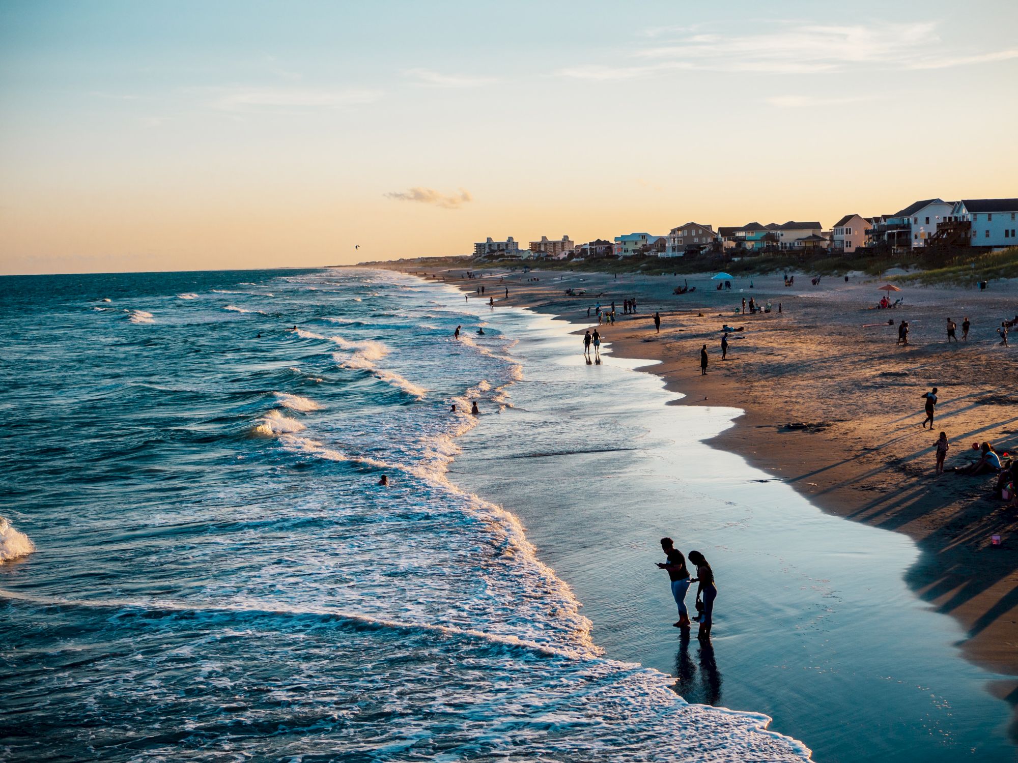 A sandy beach at sunset with people enjoying the shoreline and waves, and houses along the coast are visible in the background.