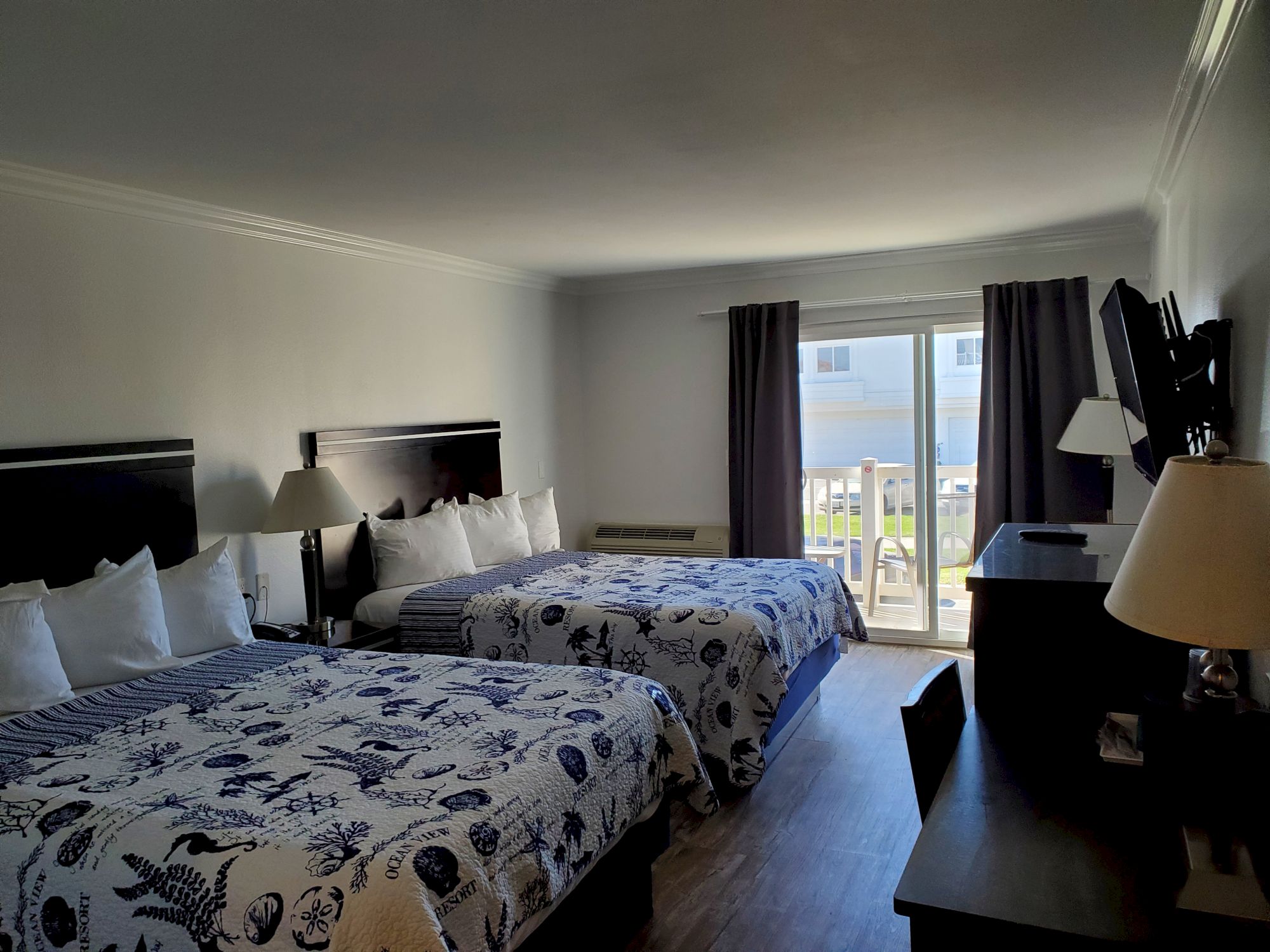 The image shows a hotel room with two double beds, a desk, a TV, and a sliding door leading to a balcony with a view.