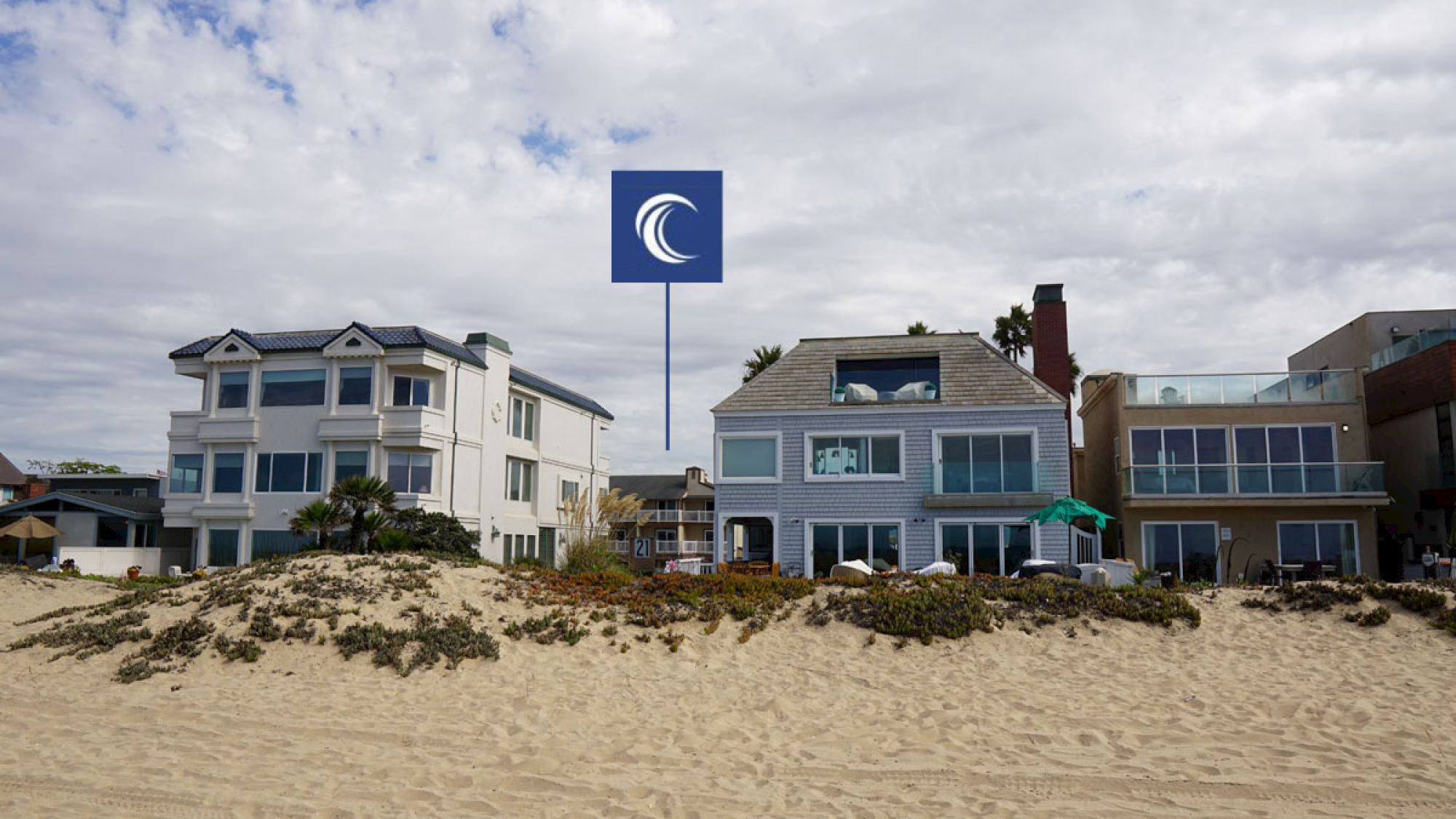 The image shows a row of beachfront houses with a logo featuring a wave above them set against a cloudy sky.