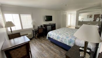 The image shows a tidy hotel room with a large bed, a desk with chairs, a flat-screen TV, lamps, and wooden flooring.