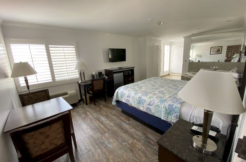 The image shows a tidy hotel room with a large bed, a desk with chairs, a flat-screen TV, lamps, and wooden flooring.