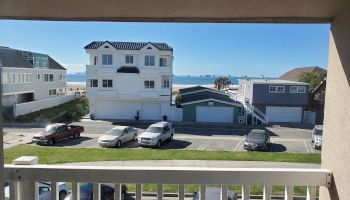 A view from a balcony overlooking parked cars, houses, and a distant beach with ships on the water under a clear blue sky.