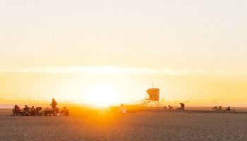 A serene beach scene at sunset with a partially visible sun, people gathered, and a lifeguard tower in the distance.