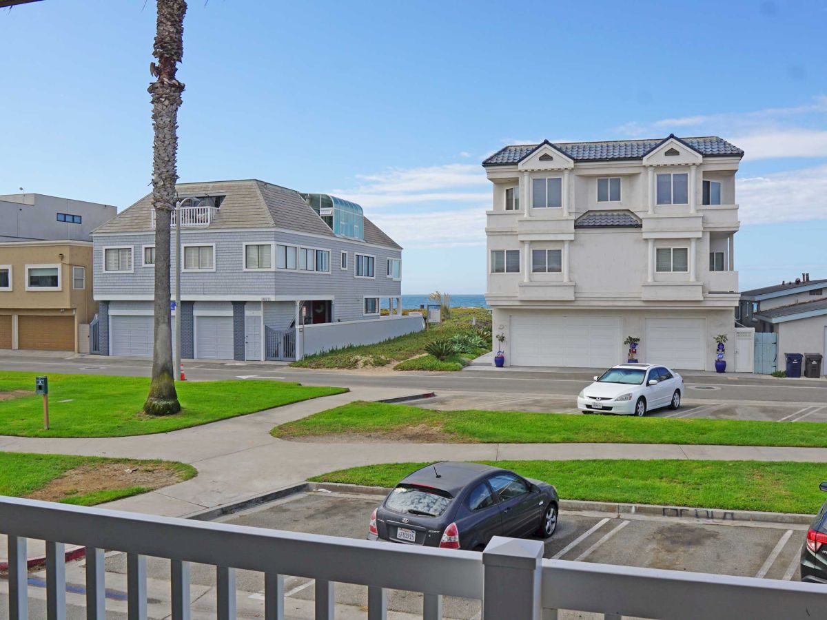 View from a balcony showing parked cars, a green lawn, palm trees, and a row of beachside homes with a glimpse of the ocean in the background.