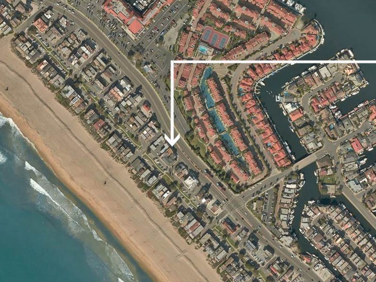 This image shows an aerial view of a coastal residential area with a beach on the left and houses, roads, and waterways on the right.