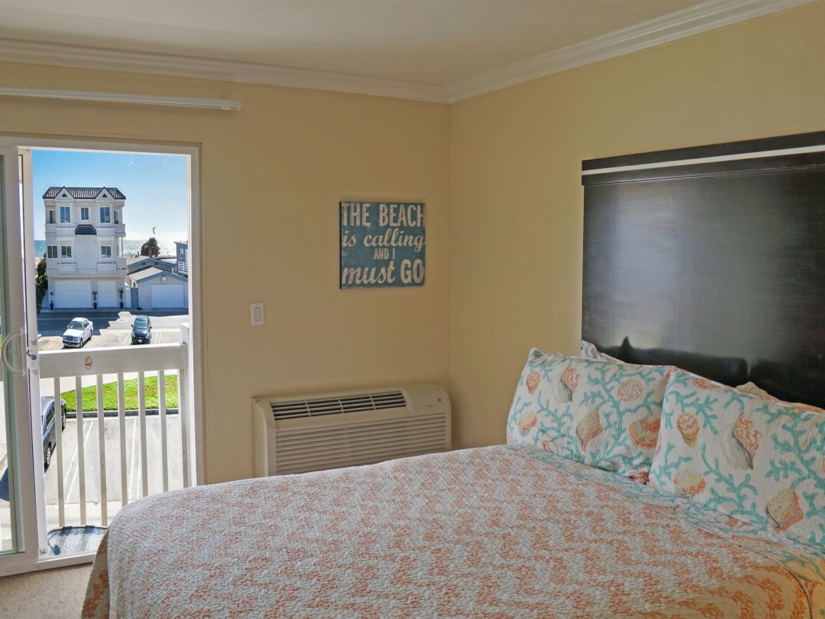 A cozy bedroom with a double bed, seashell-themed bedding, a balcony view of houses, and a sign that reads "The beach is calling and I must go."