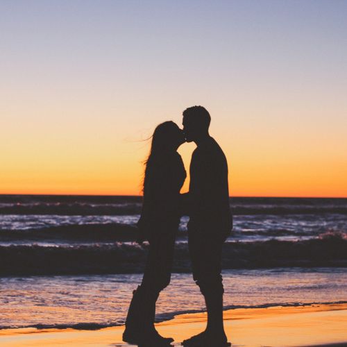 A couple is kissing at the beach during sunset, silhouetted against an orange and purple sky with waves in the background.